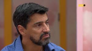 Homem de meia-idade com barba grisalha e cabelo escuro, vestindo camisa azul, em um estúdio de TV.