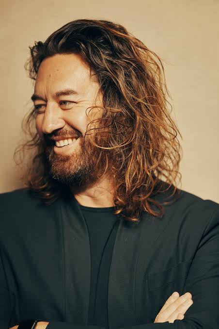 Smiling man with long hair and beard wearing a dark jacket against a plain background.