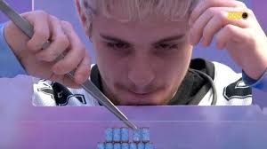 Homem com cabelo loiro e pinça manipula pequenos objetos azuis em jogo de habilidade, com fundo roxo e azul.