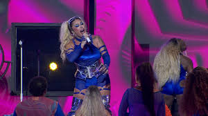 Drag queen Pabllo Vittar canta em palco com iluminação rosa e azul, vestindo traje azul brilhante e com cabelo loiro preso e…