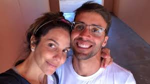 Casal sorri para a câmera em selfie. A mulher tem cabelo escuro preso com uma tiara e o homem usa óculos.