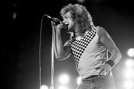 A photograph of Lou Gramm performing on stage with a microphone, surrounded by concert lighting.