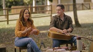 Atriz e ator sentados em cadeiras com violão e garrafa térmica em cenário rural, possivelmente de novela brasileira.