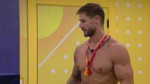 Homem musculoso com barba e medalha no pescoço em um ambiente com paredes amarelas e detalhes em branco.