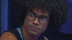 Homem com cabelo afro e óculos, participante do BBB 23, em close-up pensativo.