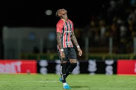 Jogador de futebol com tatuagens no corpo, vestindo uniforme listrado em preto e vermelho, com a cabeça inclinada para trás …