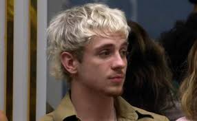 Homem com cabelo loiro e encaracolado, usando camisa bege, em um ambiente interno com outras pessoas ao fundo.