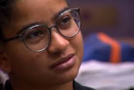 Close-up de uma jovem com óculos redondos, olhando para o lado com uma expressão pensativa. Fundo desfocado com tons de azul…