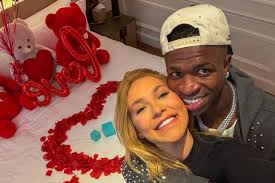 Vinícius Júnior e sua namorada em cama decorada com pétalas de rosa e ursinhos de pelúcia vermelhos.