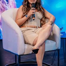 Jojo Todynho sentada em poltrona branca, segurando microfone, em evento com fundo azul e roxo.