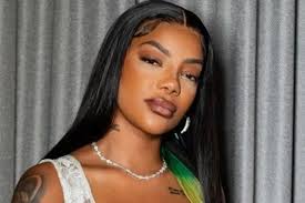 Ludmilla em close-up, com cabelo preto longo e maquiagem marcante, usando colar de pérolas e brincos.